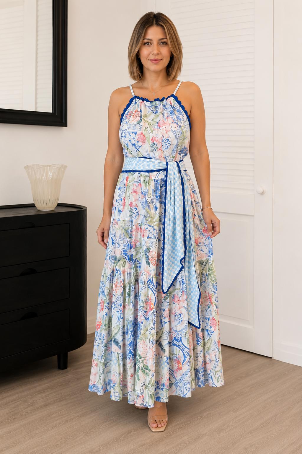 A woman modeling a sleeveless maxi dress with a blue and pink floral print, a blue patterned waist tie belt, and blue trim, standing in a room with light wood floors and white walls.