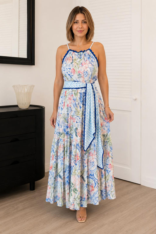 A woman modeling a sleeveless maxi dress with a blue and pink floral print, a blue patterned waist tie belt, and blue trim, standing in a room with light wood floors and white walls.
