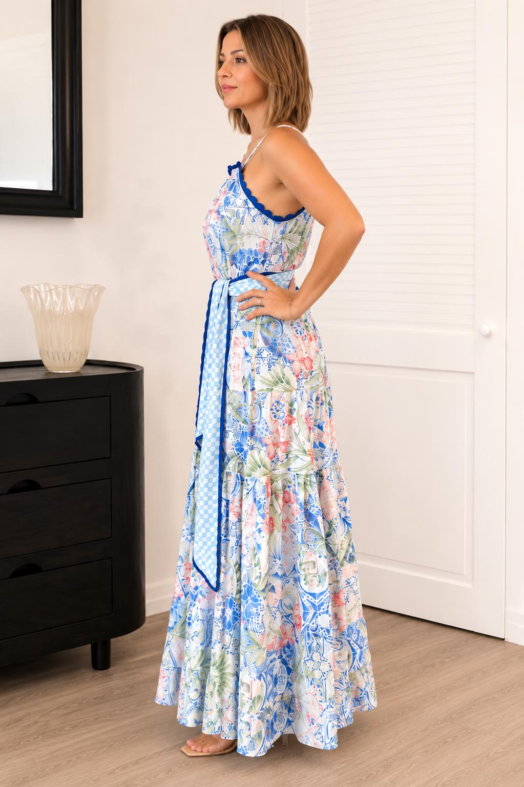 A woman modeling a sleeveless, halter-neck maxi dress with a blue and multicolor floral print, standing in a brightly lit room with a black dresser and white door in the background.