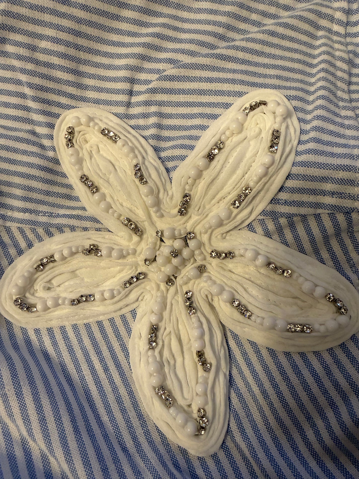 A cream-colored fabric starfish applique embellished with white beads and silver rhinestones, laid flat on a blue and white striped fabric background.