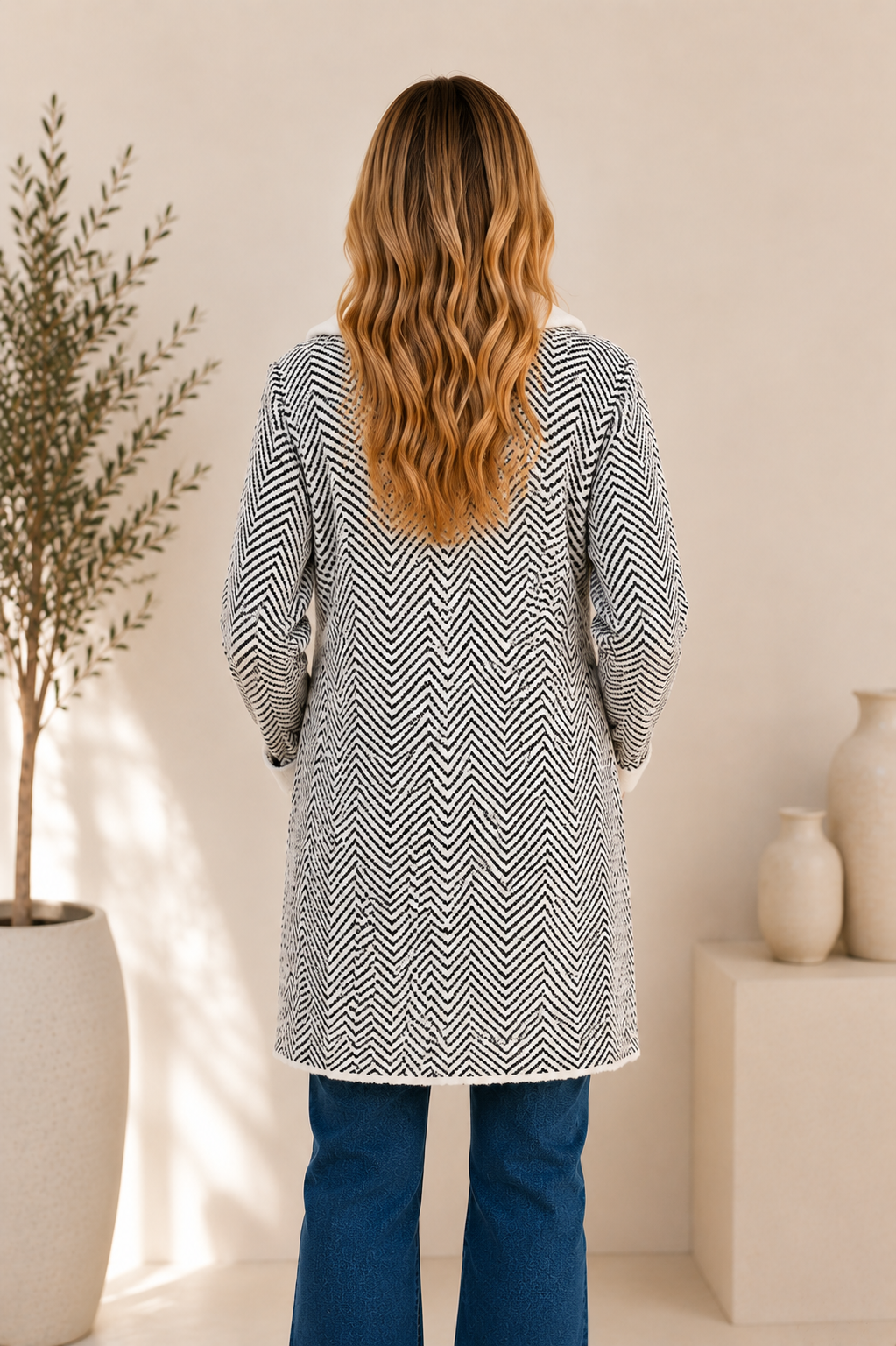 A woman viewed from behind wearing a long-sleeve tunic-style top with a black and white herringbone pattern, paired with blue jeans, in a warm-toned indoor setting with a potted plant and ceramic vases.