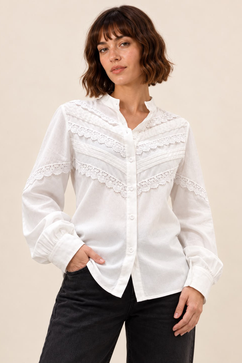A woman wearing a white long-sleeve button-up blouse with delicate lace trim across the chest and shoulders, paired with black pants, set against a solid beige studio background.