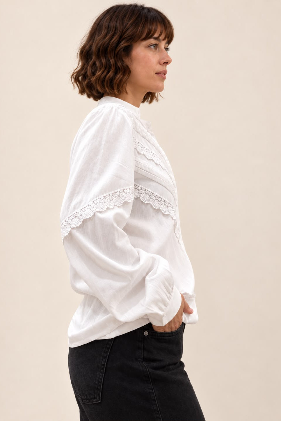 A woman in profile wearing a white long-sleeve blouse with delicate lace trim and black jeans, photographed against a plain beige studio background.