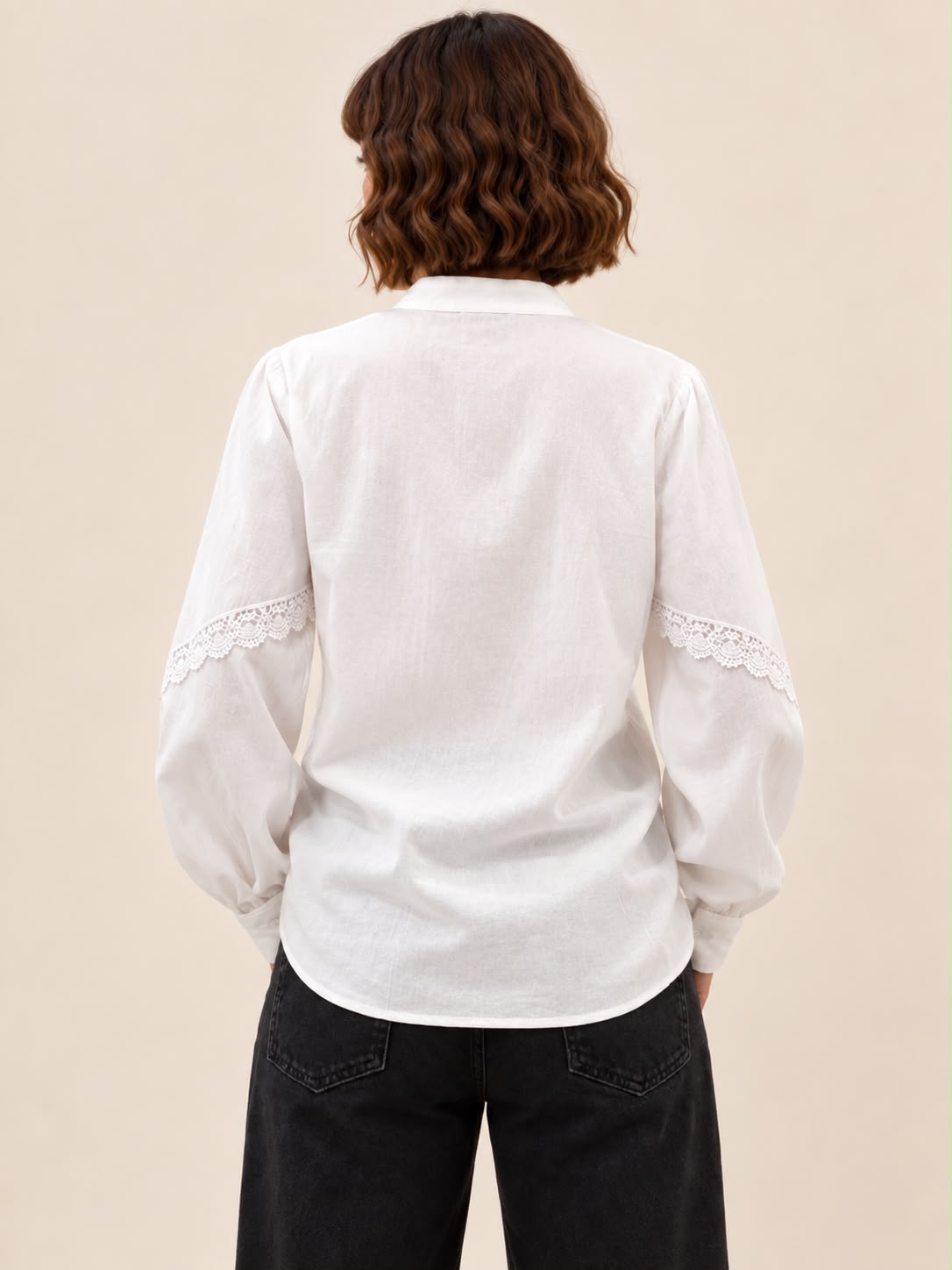 A woman viewed from behind wearing a white long-sleeve linen shirt with lace trim on the sleeves, paired with black jeans, against a solid beige background.