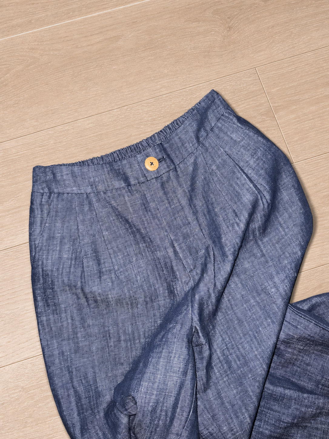 Grey-blue wide-leg trousers with an elastic waistband and wooden button, displayed on light timber floorboards.