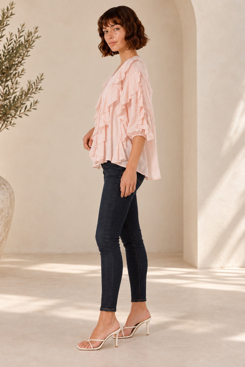 A woman stands in profile wearing a light pink ruffled blouse, dark skinny jeans, and clear high-heeled mule sandals in a bright, sunlit interior with cream walls and a large vase of greenery.